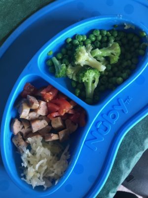 Toddler lunch in blue plate with peas, broccoli, sauerkraut, sausage, and tomatoes