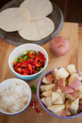 raw potatoes, onions, and bell peppers with tortillas in the background